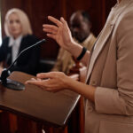 Cropped shot of young female witness in beige blazer standing by tribune with microphone and testifying in front of jury, judge and protection sideCropped shot of young female witness in beige blazer standing by tribune with microphone and testifying in front of jury, judge and protection side